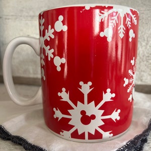 K&ouml;nnte beinhalten: Rote Keramiktasse mit einem wei&szlig;en Schneeflockenmuster und Mickey-Maus-K&ouml;pfen. Die Tasse hat einen wei&szlig;en Henkel.