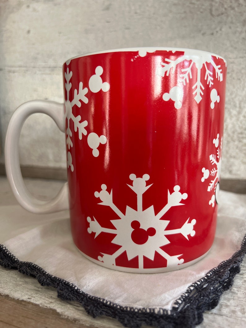 K&ouml;nnte beinhalten: Rote Keramiktasse mit einem wei&szlig;en Schneeflockenmuster und Mickey-Maus-K&ouml;pfen. Die Tasse hat einen wei&szlig;en Henkel.