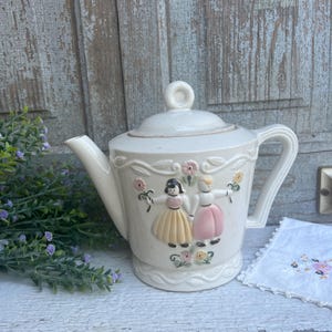 Könnte beinhalten: Weiße Teekanne aus Keramik mit Blumenmuster und zwei Mädchen, die Blumen halten. Die Teekanne hat einen Deckel mit einem Knopf oben.