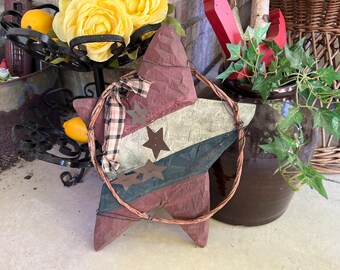 May include: A red, white and blue star-shaped wooden sign with a brown twig wreath around it. The sign has three metal stars on it. The sign is sitting on a concrete surface.