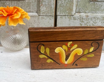 May include: A rectangular wooden box with a dark brown finish, featuring a hand-painted floral design in yellow, orange, and black on the front panel. The box has a hinged lid and a small signature mark in the bottom right corner.