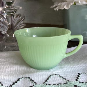 May include: A light green glass teacup featuring a ribbed, vertical texture along the lower half of the exterior. The cup has a smooth, rounded rim and a simple, angular handle extending from the side, crafted from a translucent, jade-like material.