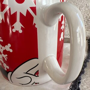 K&ouml;nnte beinhalten: Wei&szlig;e Keramiktasse mit einem roten und wei&szlig;en Schneeflockenmuster. Die Tasse zeigt eine Mickey Mouse-Silhouette und einen wei&szlig;en Henkel.