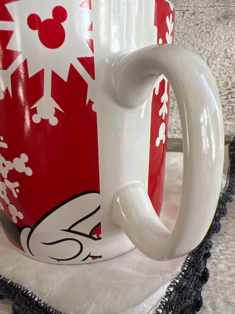 K&ouml;nnte beinhalten: Wei&szlig;e Keramiktasse mit einem roten und wei&szlig;en Schneeflockenmuster. Die Tasse zeigt eine Mickey Mouse-Silhouette und einen wei&szlig;en Henkel.