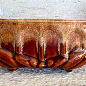May include: A rectangular brown ceramic planter with a glossy finish. The planter has a textured design with vertical arch-like patterns and sits on three small feet. The color transitions from a lighter brown at the top to a darker, richer brown at the base.