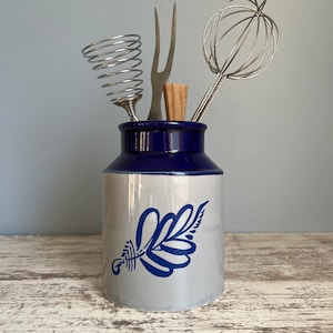 May include: A blue and white ceramic utensil holder with a leaf design. The holder has a blue rim and is filled with a whisk, a fork, and a spring-shaped tool.