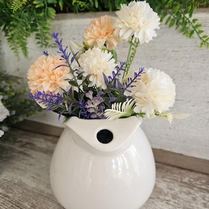 Könnte beinhalten: Eine weiße Keramikvase mit einem einzigartigen, beutelartigen Design, die ein lebendiges Arrangement künstlicher Blumen enthält. Der Strauß umfasst pfirsich- und cremefarbene Nelken sowie Zweige aus lila Lavendel und grünen Blättern. Die Vase steht auf einer Holzoberfläche.