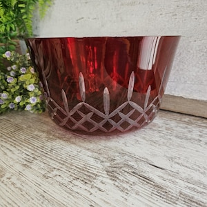 Op de afbeelding: Een robijnrode glazen kom met een diamant- en verticaal snijpatroon. De kom staat op een verweerd wit houten oppervlak. De kom is een decoratief stuk.