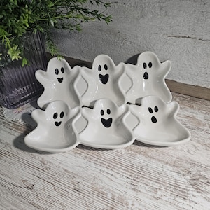Puede incluir: Seis platos blancos de cerámica con forma de fantasma y rasgos faciales negros. Los platos están dispuestos sobre una superficie de madera clara, perfectos para Halloween. Una pequeña planta en un jarrón de cristal está en el fondo.