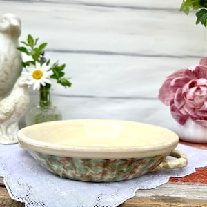 May include: A cream-colored ceramic bowl with a handle, featuring a textured green and brown exterior. The bowl sits on a white lace doily, with decorative birds and flowers in the background. A rustic, vintage-style kitchenware item.
