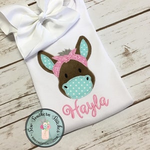 May include: White onesie with a brown donkey face with a pink bandana and a turquoise and white polka dot mask. The onesie has the name "Hayla" embroidered in pink below the donkey face. The onesie is on a white wooden surface with a white bow on top.