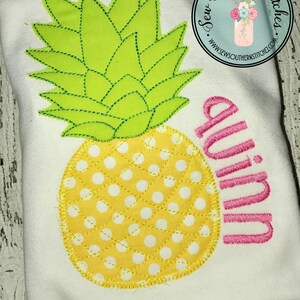 Puede incluir: Una camiseta blanca con un diseño de piña amarillo y blanco y el nombre "Quinn" bordado en rosa.