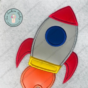 May include: A red, yellow, and blue rocket ship applique with a flame design. The rocket ship is outlined in red and has a blue window. The flame is orange and yellow.