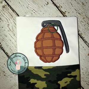 May include: A white t-shirt with a brown and gray embroidered grenade design. The shirt has a camouflage fabric bottom.