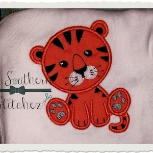 May include: Orange and black embroidered tiger applique on a white fabric background. The tiger is sitting with its paws outstretched and has a cute, cartoon-like expression.