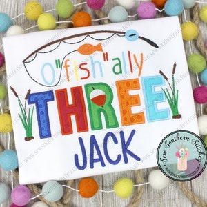 May include: A white onesie with colorful embroidered text that reads "O'fish'ally Three Jack". The text is surrounded by a fishing rod, a fish, and reeds.