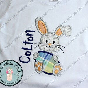 May include: White t-shirt with an embroidered Easter bunny holding a plaid egg. The bunny is white with orange and grey accents. The name "Colton" is embroidered in navy blue. The shirt has a round neck and short sleeves.