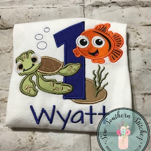 May include: A white onesie with a blue number one, a green turtle, and an orange fish. The onesie is personalised with the name Wyatt. The text "Sew Southern Stitches" is in the bottom right corner.