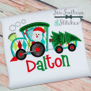May include: White t-shirt featuring a colorful Christmas train design. The train has Santa Claus in the engine and a Christmas tree in the car. The name "Dalton" is written in red and green below the train.