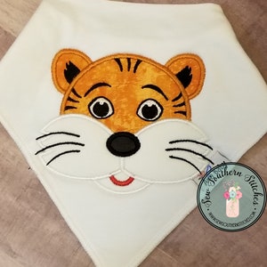 May include: White bib with an embroidered tiger face. The tiger has orange fur, black stripes, black eyes, a black nose, and a red mouth.
