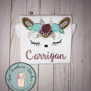 Pode incluir: Camiseta branca com um rosto de veado marrom bordado. O veado tem uma coroa floral com flores rosa, vermelhas e azuis. O nome "Carrigan" está bordado em rosa abaixo do veado. A camiseta tem um logotipo Sew Southern Stitches no canto inferior esquerdo.