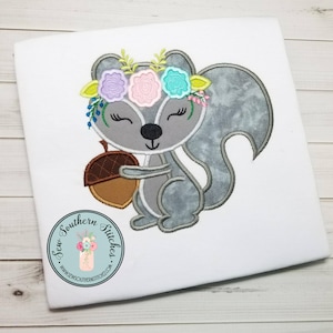 May include: Embroidered design of a gray squirrel holding an acorn. The squirrel is wearing a flower crown with pink, blue, and yellow flowers. The design is on a white t-shirt.