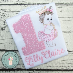 May include: White shirt with a pink number one applique and a princess design. The princess has a crown, a tutu, and a pearl necklace. The name "Lilly Claire" is embroidered in pink.