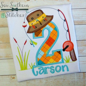 May include: A white t-shirt with an embroidered design of a fishing hat, a fishing rod, a number 2, and the name Carson. The number 2 is made of orange, blue, and yellow fabric. The design is surrounded by green grass and red reeds.