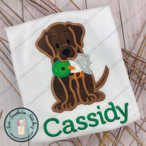 May include: A white t-shirt with an embroidered brown labrador retriever holding a green and white duck. The name "Cassidy" is embroidered in green below the dog.
