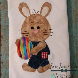 May include: A white fabric item featuring an appliqued Easter bunny holding a colorful striped egg. The bunny is brown with cream-colored ears and wears denim overalls with a red and white checkered pocket. The text "Sew Southern Stitchez" is at the bottom.