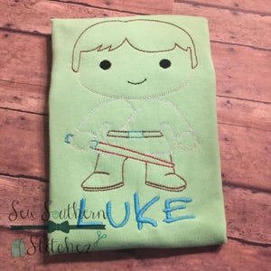 Op de afbeelding: Een lichtgroen T-shirt met een geborduurd beeld van een cartoonfiguur dat een lightsaber vasthoudt. Het figuur draagt een bruine riem en bruine laarzen. De tekst "LUKE" is in blauw onder het figuur geborduurd.
