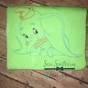 Puede incluir: Diseño bordado de un elefante azul claro con un sombrero rojo y una bufanda roja y blanca en una camiseta verde. El diseño está cosido con hilo azul. La camiseta tiene el texto "Sew Southern Stitchez" en negro con una corbata de lazo y tijeras.