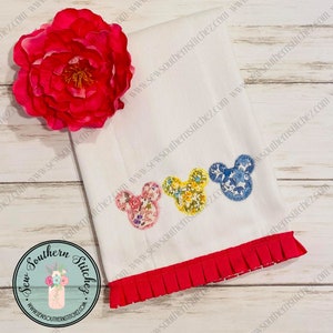 May include: A white kitchen towel with a red ruffled edge. Three embroidered Mickey Mouse heads are on the towel. Each head is made of different floral patterned fabric. The heads are in a row, with the pink head on the left, the yellow head in the middle, and the blue head on the right.