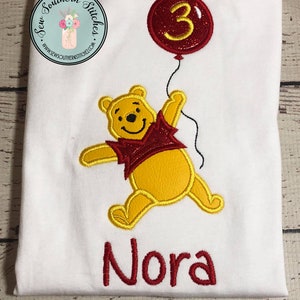 May include: White t-shirt with a red embroidered balloon with the number 3, a yellow embroidered Winnie the Pooh character holding the balloon, and the name "Nora" embroidered in red.