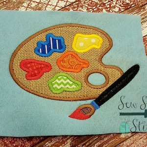 May include: A brown burlap paint palette with colourful paint swatches and a paintbrush. The palette has a hole in the centre and is surrounded by a brown outline. The paintbrush is black with a blue handle and a red tip. The background is a light blue felt.