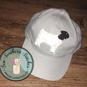 Peut inclure: Casquette de baseball grise avec une applique de chèvre blanche et noire. La casquette a une visière incurvée et un logo cousu qui indique "Sew Southern Stitches. www.sewsouthernstitches.com".