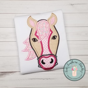 May include: Embroidered horse head design in pink, brown, and white on a white t-shirt. The horse has a pink mane and a pink and white checkered noseband.