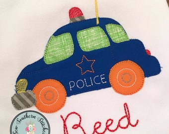 May include: A blue and green police car applique with orange wheels and a red light on top. The car has a white star and the word "POLICE" embroidered on it. The name "Reed" is embroidered in red below the car.