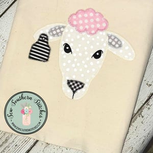 May include: White felt applique of a sheep's head with black and white polka dots, black and white checkered nose, and pink felt flower with pink polka dots on top. The applique is on a beige fabric background.