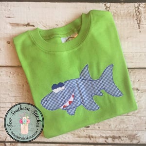 Op de afbeelding: Een limoengroen T-shirt met een blauwe en witte gingham-haaiapplicatie. De haai heeft een tandenrij en één oog.