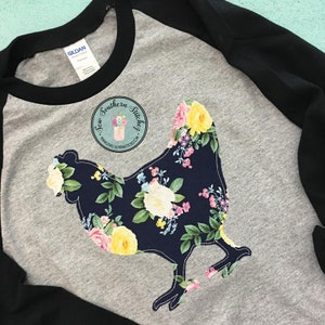 May include: A grey and black baseball tee shirt with a floral print chicken silhouette. The shirt has the text "Sew Southern Stitches" with a website address.