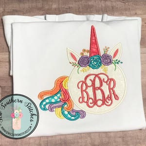 May include: White embroidered unicorn with a rainbow mane and tail. The unicorn has a pink horn and a floral crown with purple, pink, and blue flowers. The unicorn's body is a circle with the monogram "RBR" in the center.