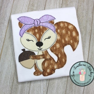 May include: A brown and white spotted squirrel with a purple polka dot bow holding a brown acorn. The squirrel is embroidered on a white t-shirt. The text "Sew Southern Stitches" is in a circle on the bottom right corner of the image.