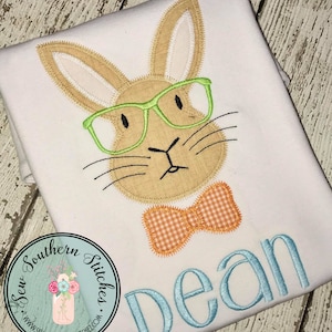 May include: A white t-shirt with an embroidered brown bunny wearing green glasses and an orange bow tie. The name "Dean" is embroidered in blue below the bunny.