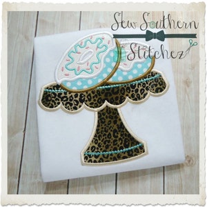 Puede incluir: Un soporte para pastel blanco con un diseño de estampado de leopardo, coronado con dos donuts glaseados con chispas. El soporte tiene un borde festoneado y una base de pedestal. El texto "Sew Southern Stitchez" es visible en la esquina superior derecha de la imagen.