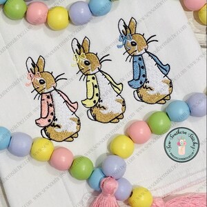 May include: Three embroidered Peter Rabbit characters on a white fabric background. The rabbits are wearing different colored jackets: pink, yellow, and blue. The background is decorated with a garland of pastel colored wooden beads.