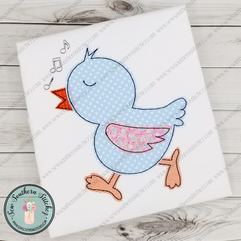 K&ouml;nnte beinhalten: Ein blauer und rosa Applikationsvogel mit einem Blumenmuster auf seinem Fl&uuml;gel. Der Vogel singt mit Noten &uuml;ber seinem Kopf. Die Applikation befindet sich auf einem wei&szlig;en Hintergrund.