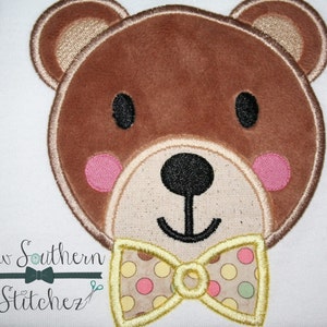 May include: Embroidered brown bear face with pink cheeks and a yellow bow tie with a polka dot pattern. The bow tie has yellow, pink, green, and white polka dots.