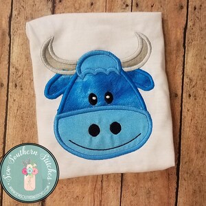 May include: A blue embroidered bull applique with white horns on a white t-shirt. The bull has a smiling face with black eyes and a black nose.