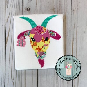 May include: A white t-shirt with a colorful patchwork goat applique. The goat has a pink nose, black eyes, and a pink tag with the letter "H" on it. The t-shirt is decorated with a Sew Southern Stitches logo.